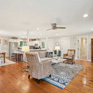A bright, open-concept living and dining area with wooden floors, white walls, a blue rug, wicker chairs, a kitchen island, and a TV mounted on a wall.