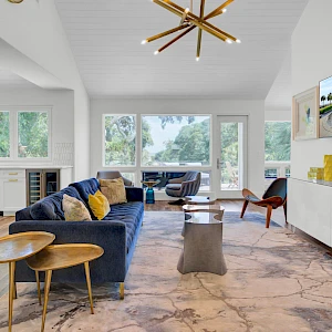 A bright, modern living room with a blue sofa, mid-century chairs, a wall-mounted TV, large windows, and a marble floor, airy and stylish.