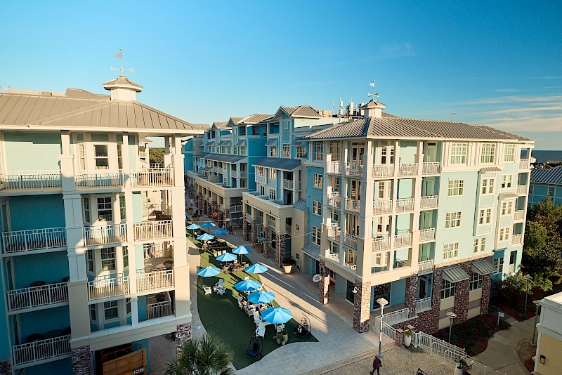 Cozy oceanfront condo complex with pastel blue buildings, balconies, and a courtyard lined with blue umbrellas and seating along a sandy path.