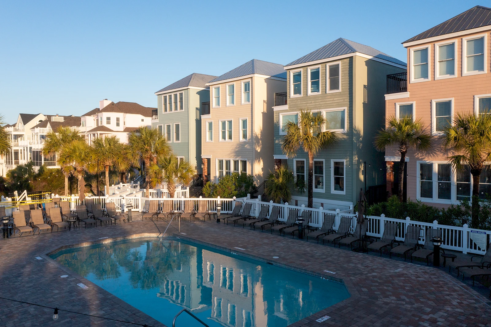 A residential community with colorful townhouses surrounding a rectangular swimming pool, lounge chairs, and palm trees in a sunny courtyard.