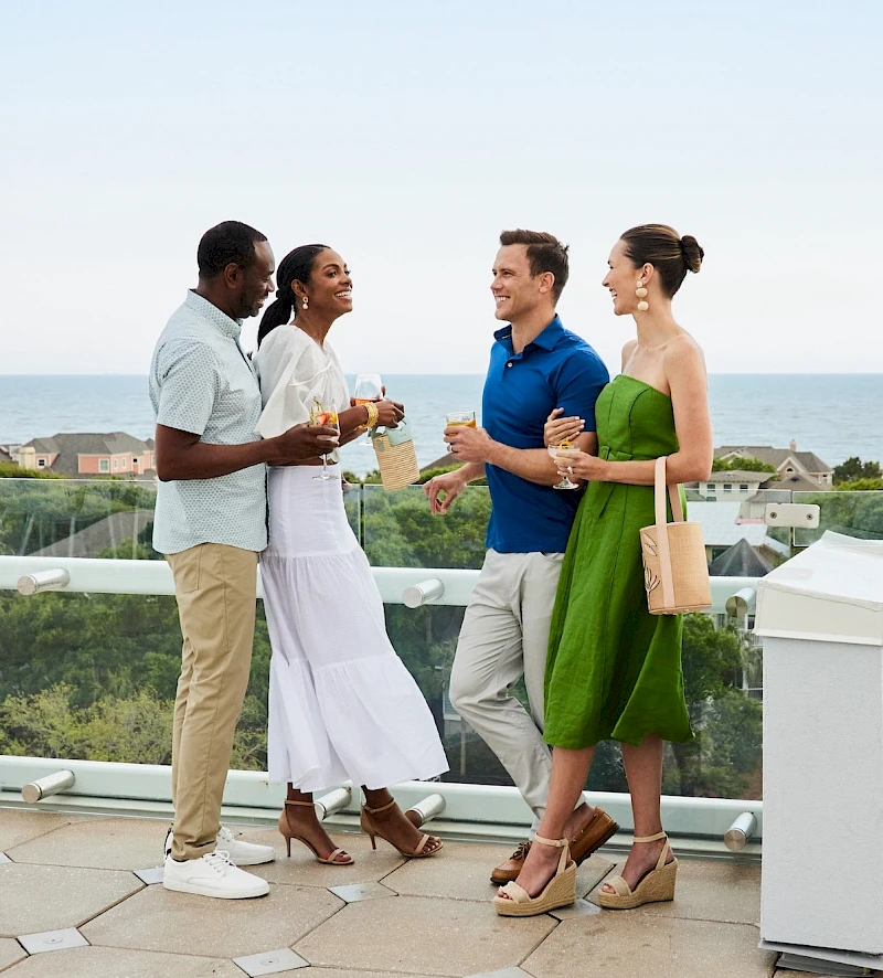 A group of four friends chatting and toasting drinks on a balcony with a sea view, dressed in smart casual attire.
