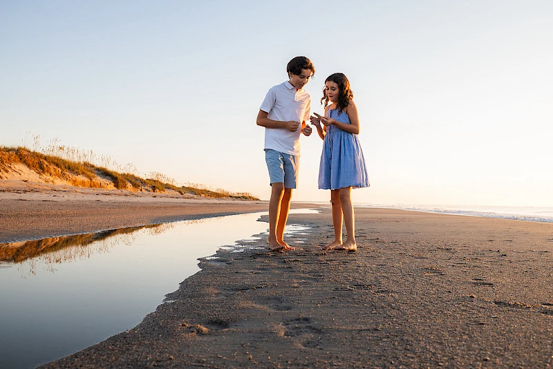 Two people stand on a sandy beach near a shallow tidal pool, chatting and smiling as the sun sets, enjoying a calm, breezy moment.