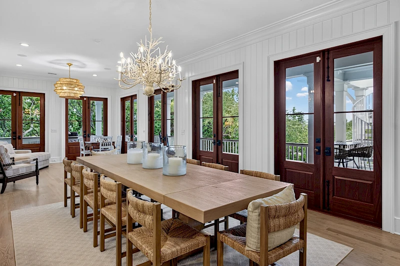 A bright dining room with a long wooden table, woven chairs, glass doors, chandeliers, and neutral decor, opening to an outdoor view.