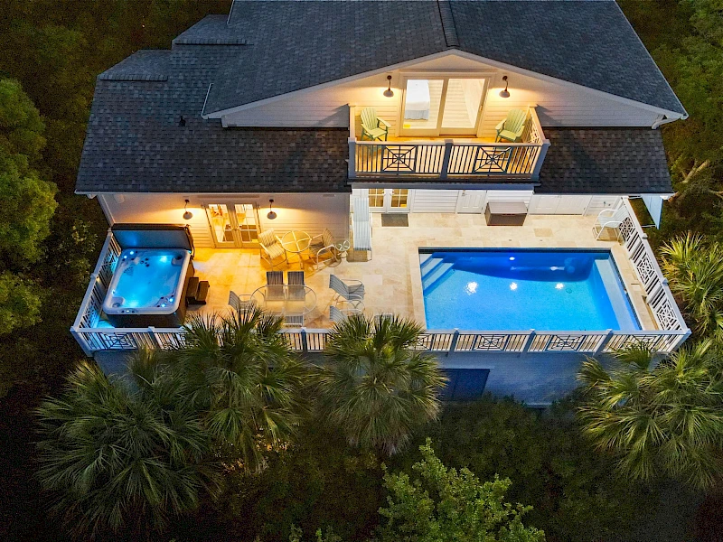 Luxurious two-story villa with a illuminated outdoor lounge, a bright pool, a hot tub on a balcony, and lush surrounding palm trees at dusk.
