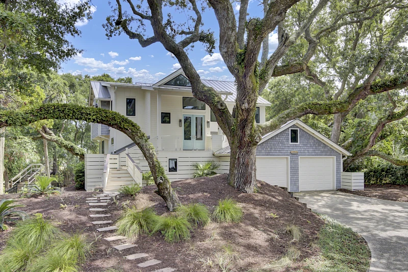 A two-story light-gray house with a garage, mature trees arching over the front yard, stone steps, and a sloped landscaped driveway.