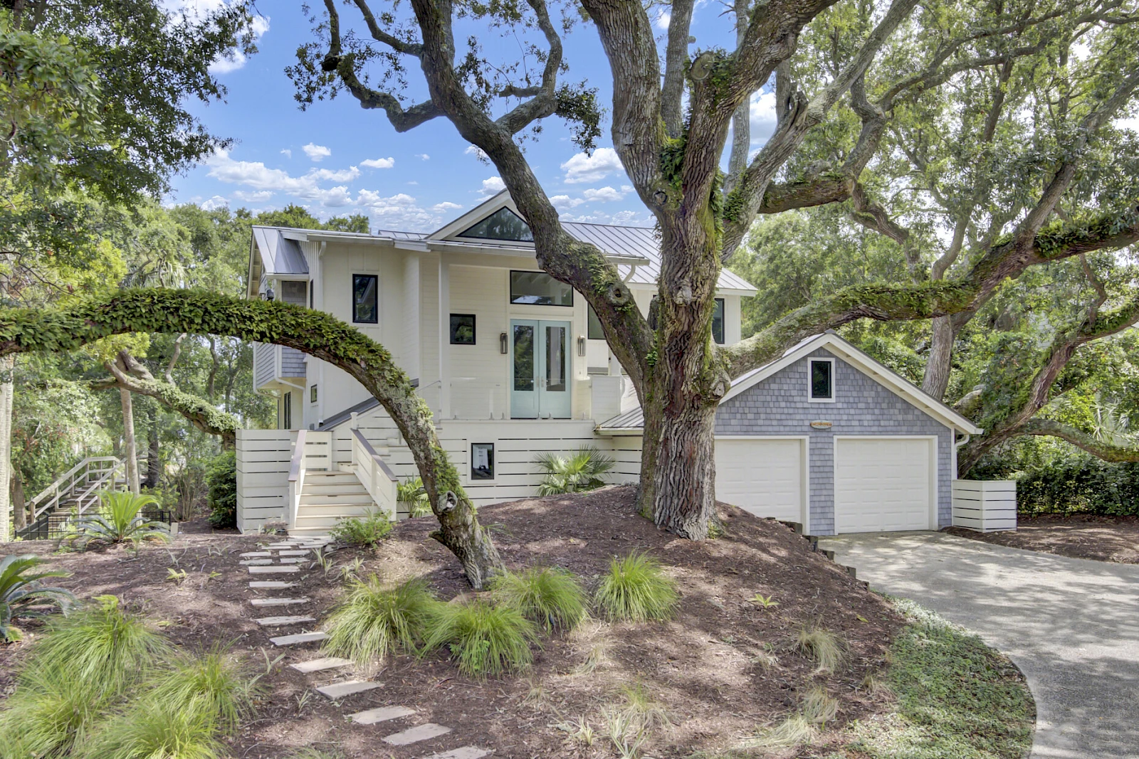 A white two-story house with a detached two-car garage, surrounded by mature trees and a sloped landscape with stepping stones.