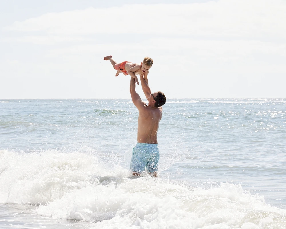 A man in blue swim trunks lifts a small orange dog above his head in the ocean waves at the shore, sunny seaside scene.