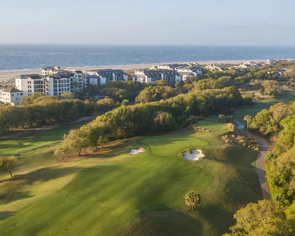 A coastal golf course with green fairways, sand bunkers, trees, and a residential area in the background along the shore.