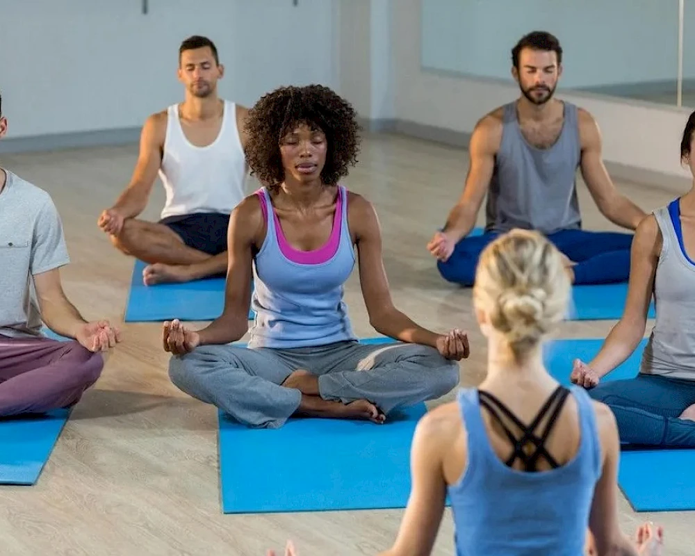 People meditating in a yoga class: adults on mats in a calm, seated pose with eyes closed and a trainer guiding at the front.