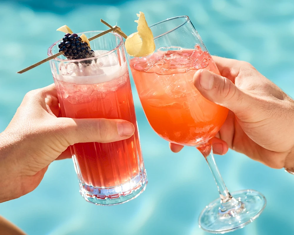Two colorful tropical cocktails clink in a poolside toast, garnished with fruit, sunshine, and relaxed vacation vibes.