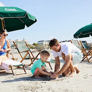 A family builds a sandcastle on the beach under green umbrellas, with a woman watching, a man helping a child, and a sunny coastline in the background.