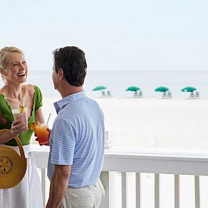 Two people chat and smile on a beachside balcony, holding drinks and a sun hat, with the ocean and umbrellas in the background.