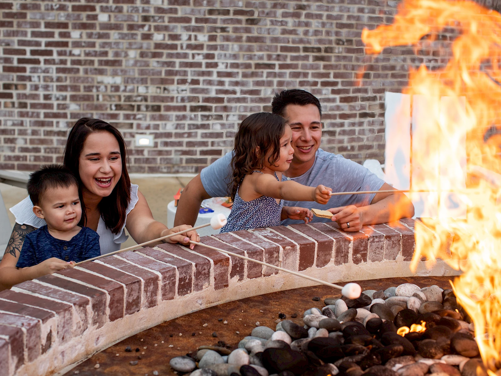 A family of four watches a bright fire pit flare up in a circular brick surround, smiling and waving hands near the flames.