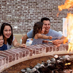A family of four watches a bright fire pit flare up in a circular brick surround, smiling and waving hands near the flames.