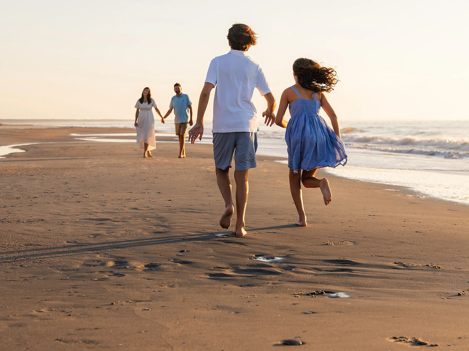 A family runs hand-in-hand along a sunny beach, footprints trailing in the wet sand as the ocean sparkles nearby.