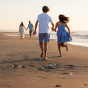 A family runs hand-in-hand along a sunny beach, footprints trailing in the wet sand as the ocean sparkles nearby.