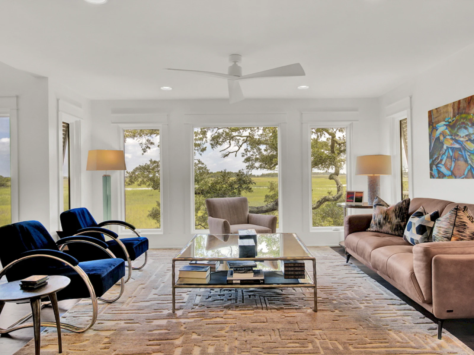 A modern living room with large windows, a beige sofa, blue armchairs, a glass coffee table, wooden floors, and greenery visible outside, bright and airy.
