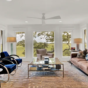 A modern living room with large windows, a beige sofa, blue armchairs, a glass coffee table, wooden floors, and greenery visible outside, bright and airy.