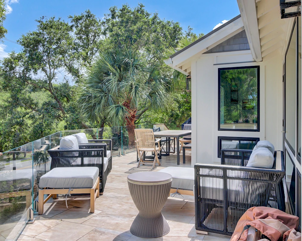 A sunny balcony or deck with outdoor seating, a dining table, potted plants, and a view of trees in a suburban backyard, bright and inviting.