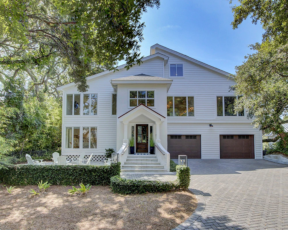 A large white two-story house with a gabled roof, two garages, a centered front door, and surrounding green trees along a paved driveway, bright and inviting.