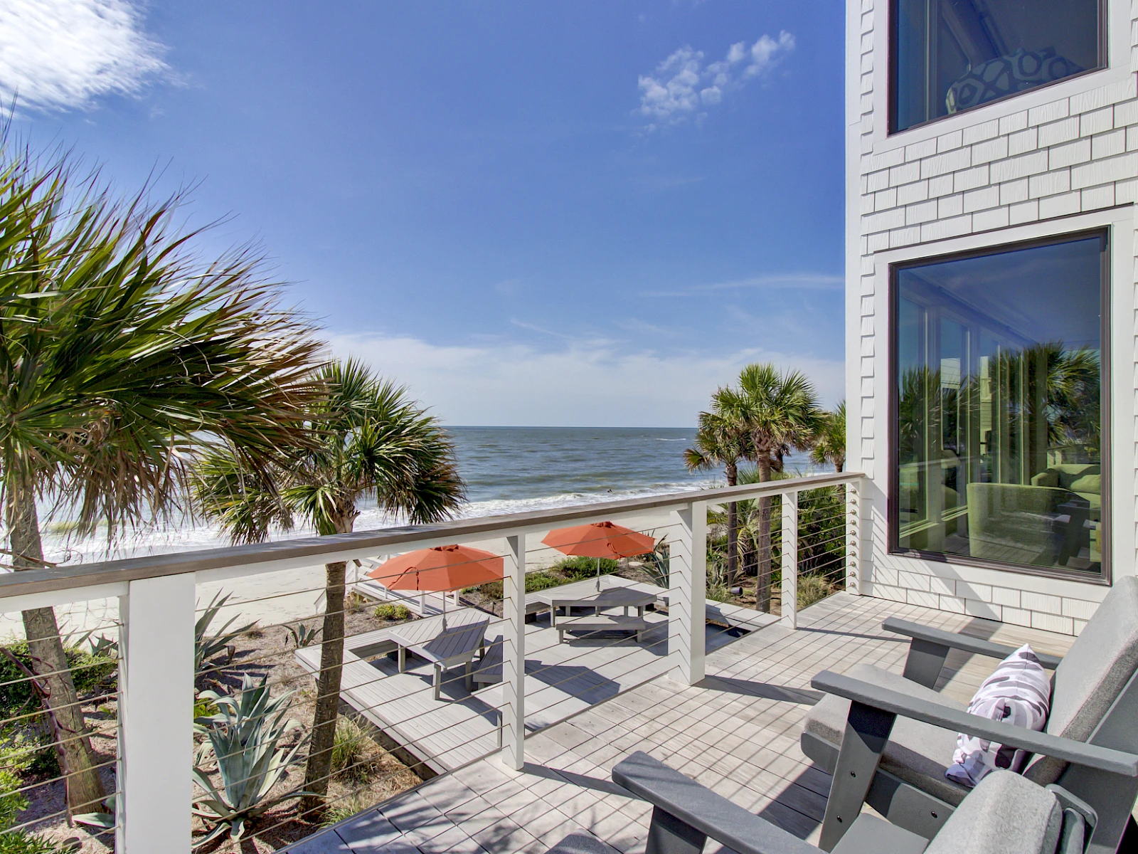 A bright beach balcony with white railings, palm trees, and umbrellas, overlooking the ocean; cozy chairs invite sea breeze and relaxing views.