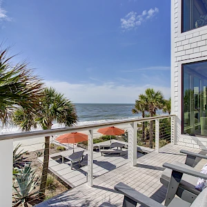 A bright beach balcony with white railings, palm trees, and umbrellas, overlooking the ocean; cozy chairs invite sea breeze and relaxing views.