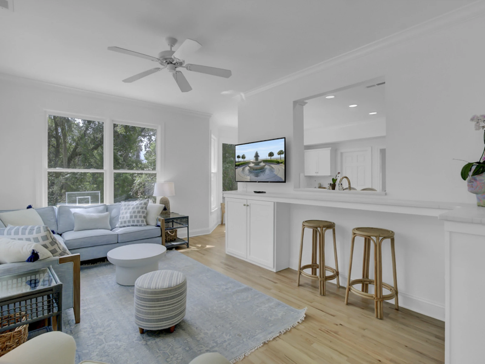 Bright, airy living area with white walls, a light wood floor, sofa seating, a ceiling fan, large window, and a breakfast bar with stools.