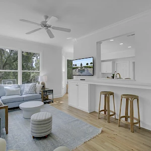 Bright, airy living area with white walls, a light wood floor, sofa seating, a ceiling fan, large window, and a breakfast bar with stools.