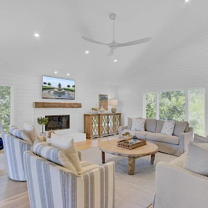 Bright, airy living room with white walls, large windows, soft gray sofas, striped chairs, stone fireplace, and a hanging egg chair by the window.