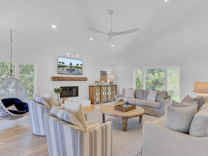 Bright, airy living room with white walls, large windows, soft gray sofas, striped chairs, stone fireplace, and a hanging egg chair by the window.