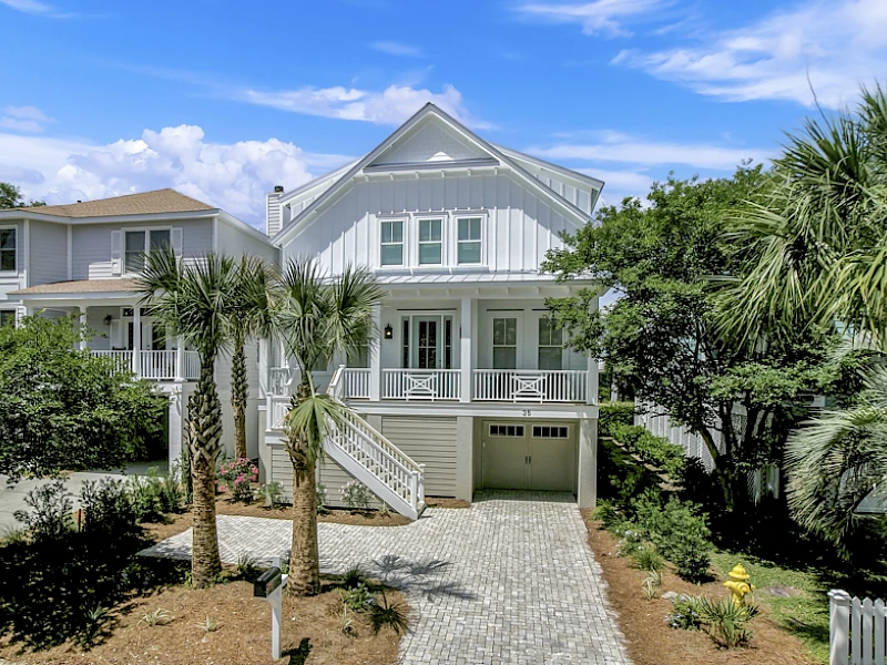 A bright coastal-style house with a second-floor balcony, palm trees, a paved driveway, and a two-car garage fronting a sunny yard, overall tropical and inviting.