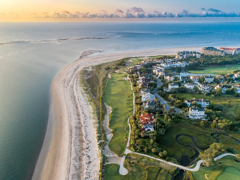 Aerial view of a coastal resort with a sandy beach, ocean, and a row of houses and golf courses inland, connected by winding paths and greens, serene seascape.