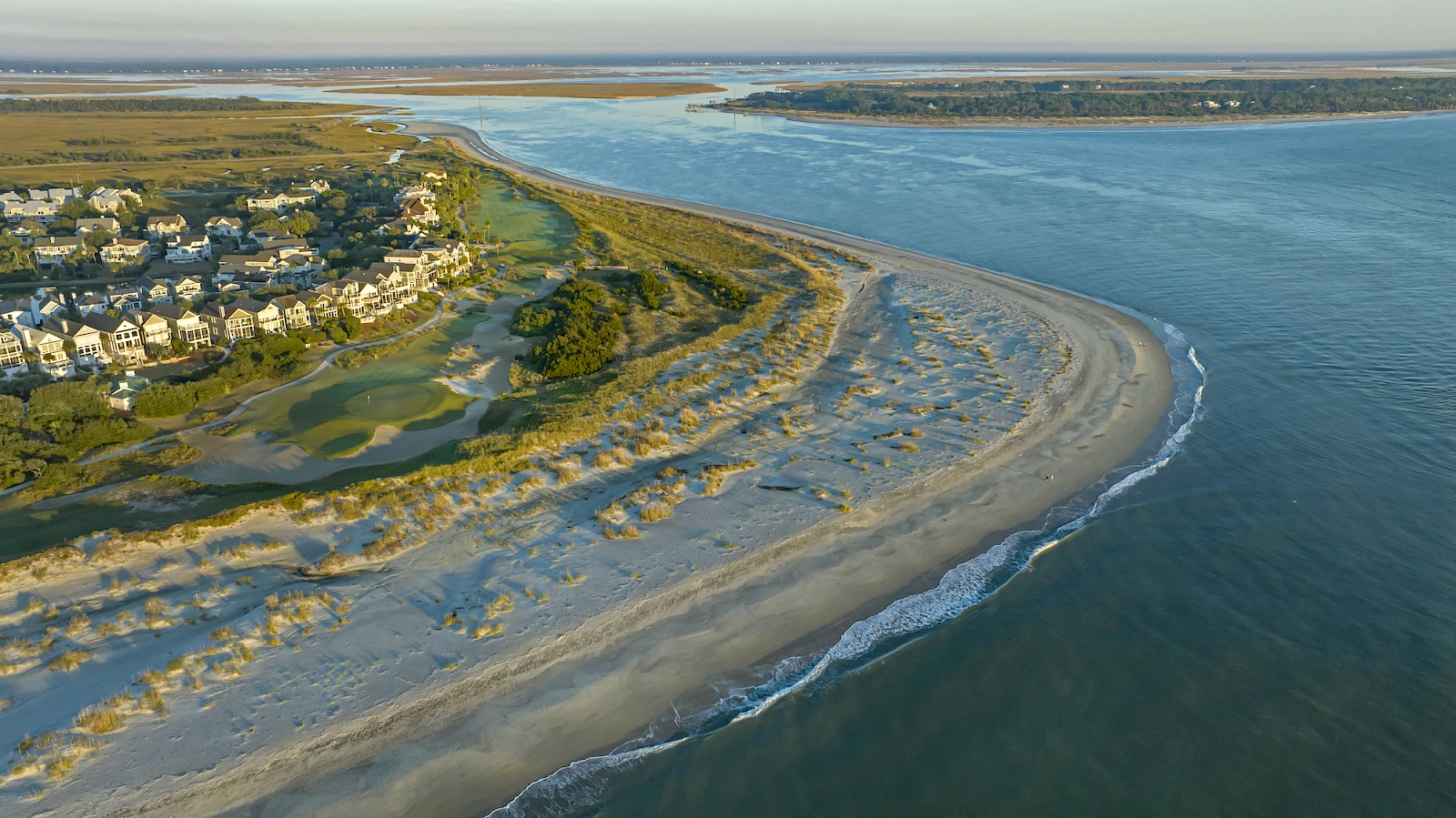 An aerial view of a sandy coastal inlet with a curved barrier beach, small houses on the left, grassy dunes, and calm blue water on the right.