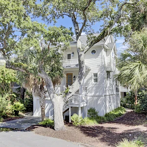 A light gray, two-story townhouse with white trim, front steps, surrounding trees, shrubs, and a small patch of mulch in the yard.