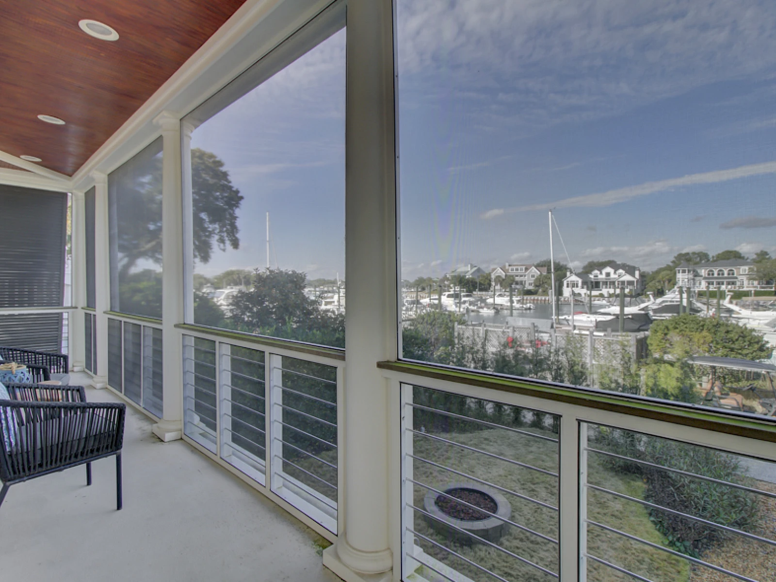 Balcony with two chairs overlooks a marina and residential houses, clear blue sky, calm water, and distant boats in a sunny coastal scene.