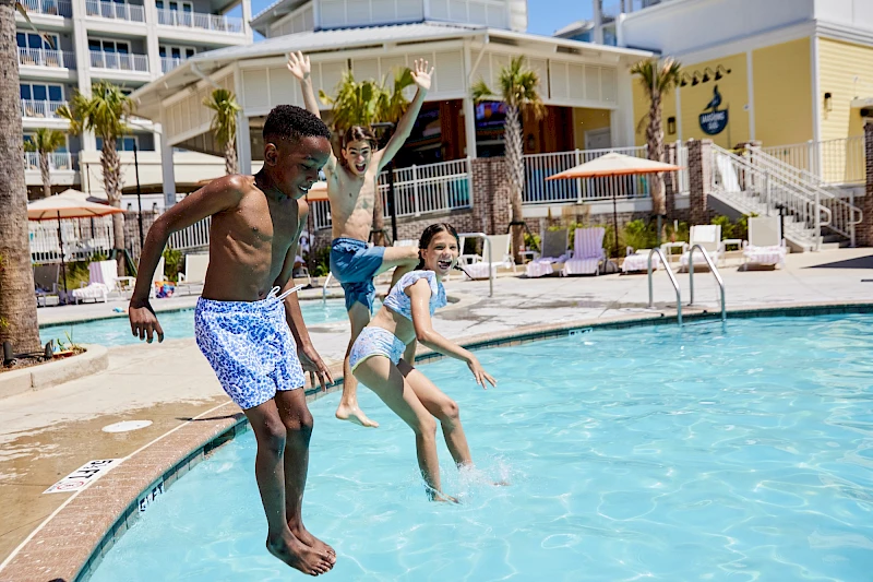 Two kids jump into a sunny pool at a resort, leaving splashes behind as others lounge and buildings frame the scene.