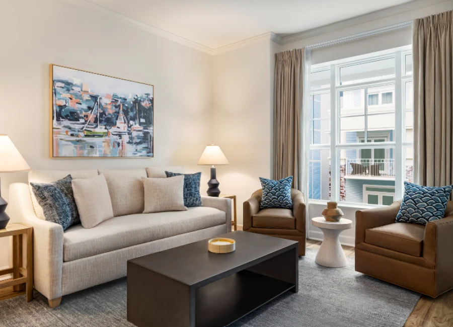 A cozy living room with a light beige sofa, two brown armchairs, a dark coffee table, corner lamps, a painting, and a large window with beige curtains.
