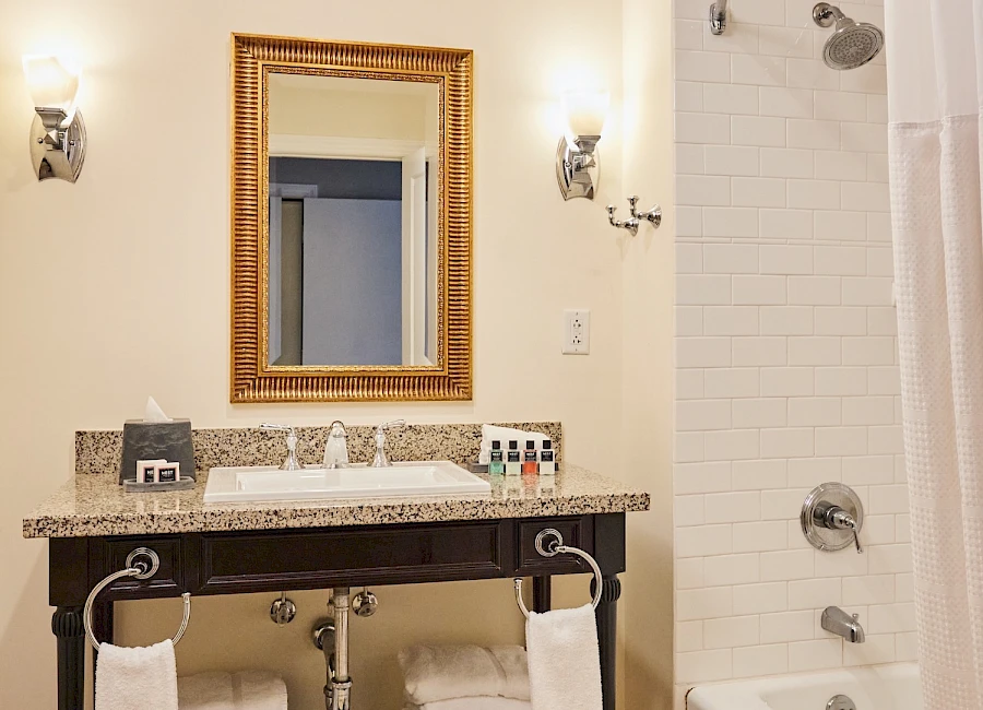 A stylish bathroom with a granite-topped vanity, a gold-framed mirror, stacked towels on a lower shelf, and a white tiled shower/bath combo.