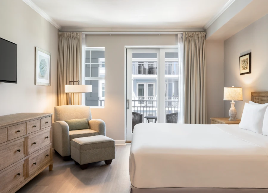 A bright hotel room with a large bed, a dresser, a cozy armchair and ottoman, a floor lamp, wall art, a TV, and a balcony view through glass doors.