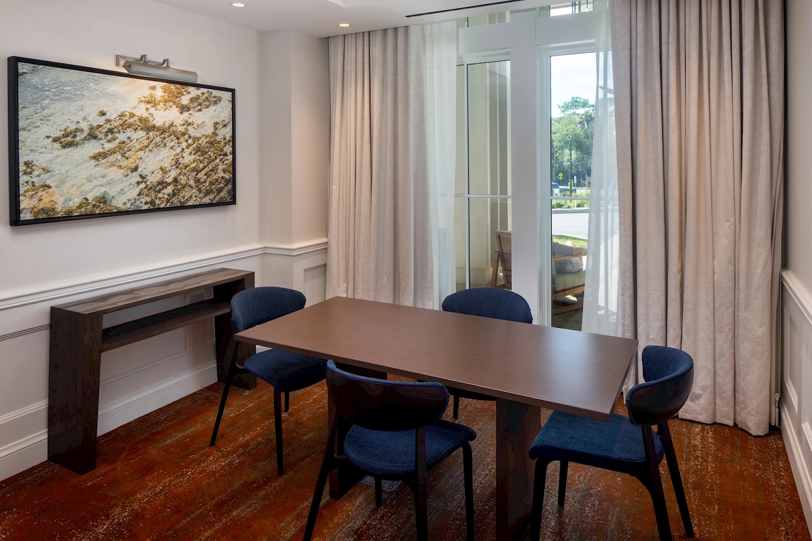 A small dining area with a brown table and six blue chairs, a wall-mounted art piece, a console table, and large windows with light curtains.