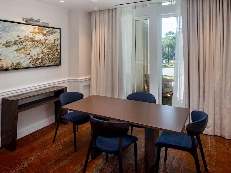 A small dining area with a brown table and six blue chairs, a wall-mounted art piece, a console table, and large windows with light curtains.