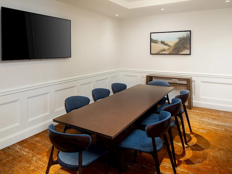 A conference room with a long dark table, blue upholstered chairs, white wainscoted walls, a wall-mounted TV, and a framed landscape photo.