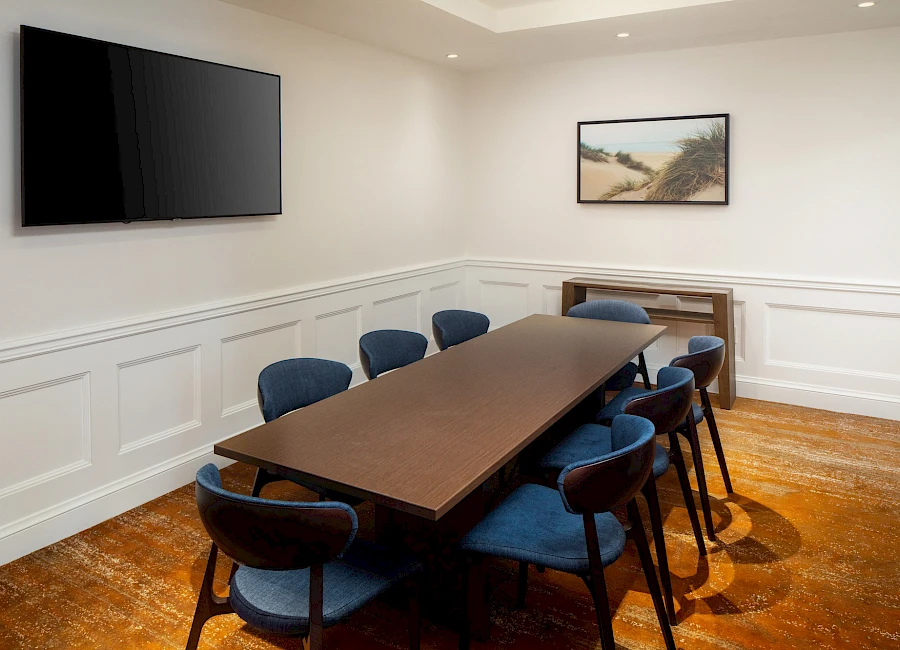 A conference room with a long dark table, blue upholstered chairs, white wainscoted walls, a wall-mounted TV, and a framed landscape photo.