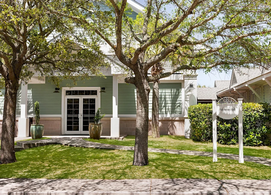 A pastel green house with a shaded front porch, two large trees, a lawn, potted plants near the entrance, and a white signpost by the yard.