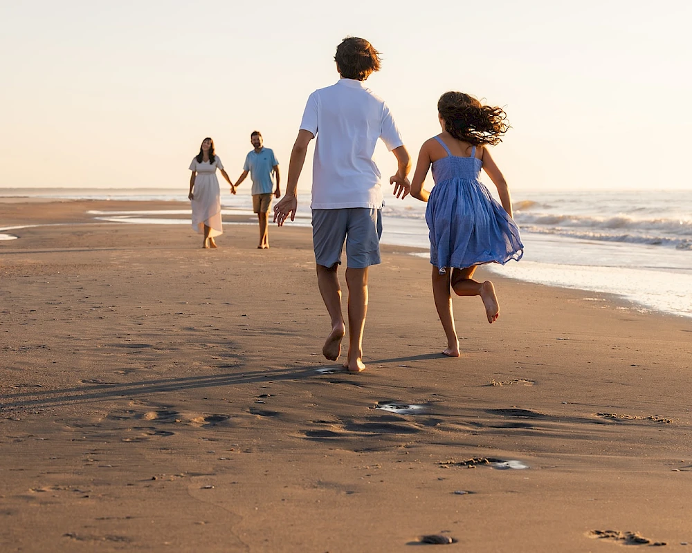 A family of four runs along a sandy beach at sunset, the kids in blue dresses, waves lapping the shore, footprints trailing behind them.