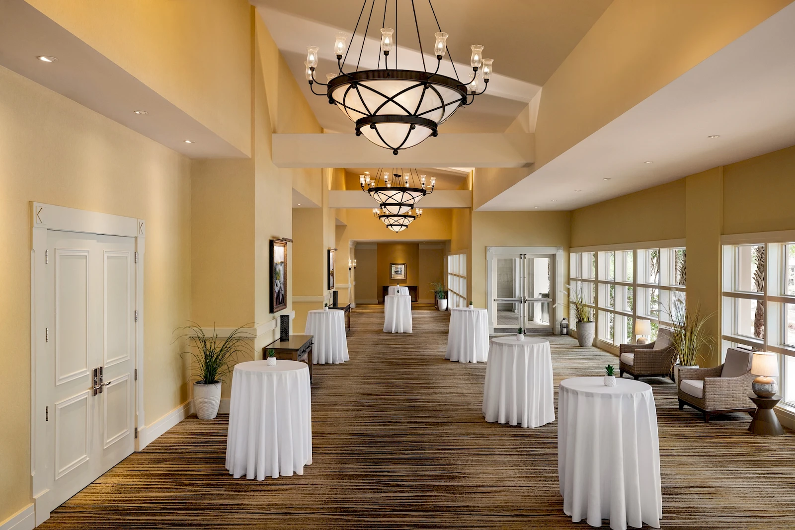 A spacious event hall with high ceilings, chandeliers, and tall cocktail tables draped in white cloth.