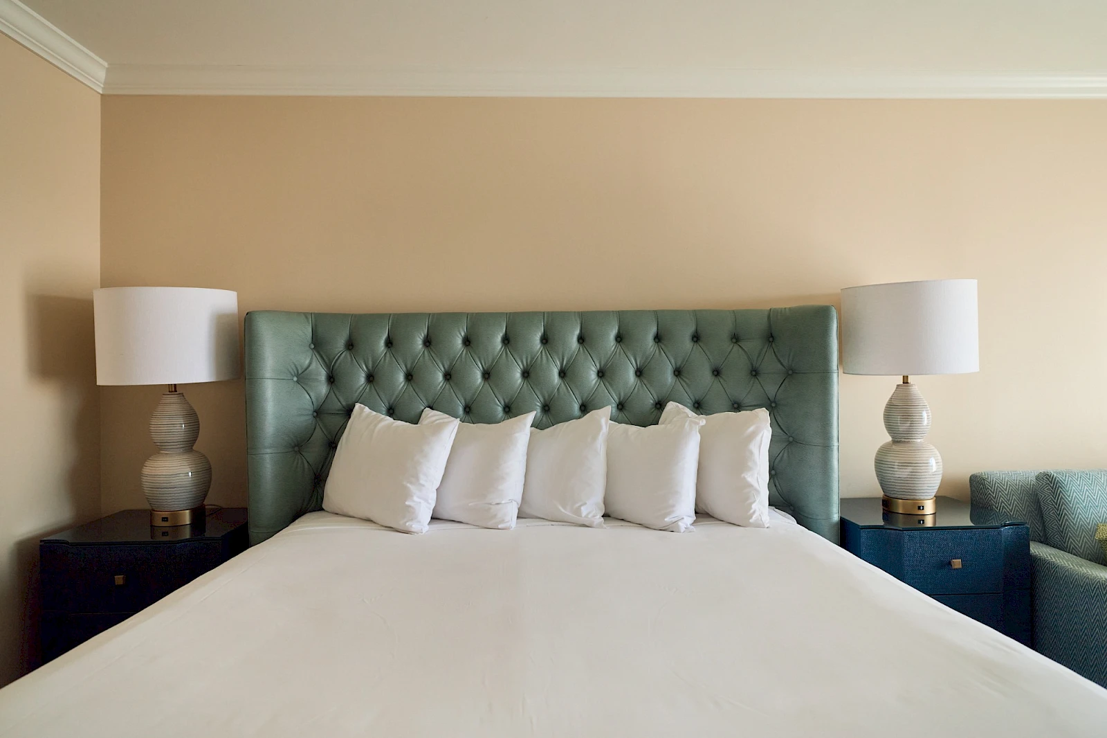 A neatly made bed with a tufted green headboard, six white pillows, cream walls, and matching nightstands with lamps on either side.