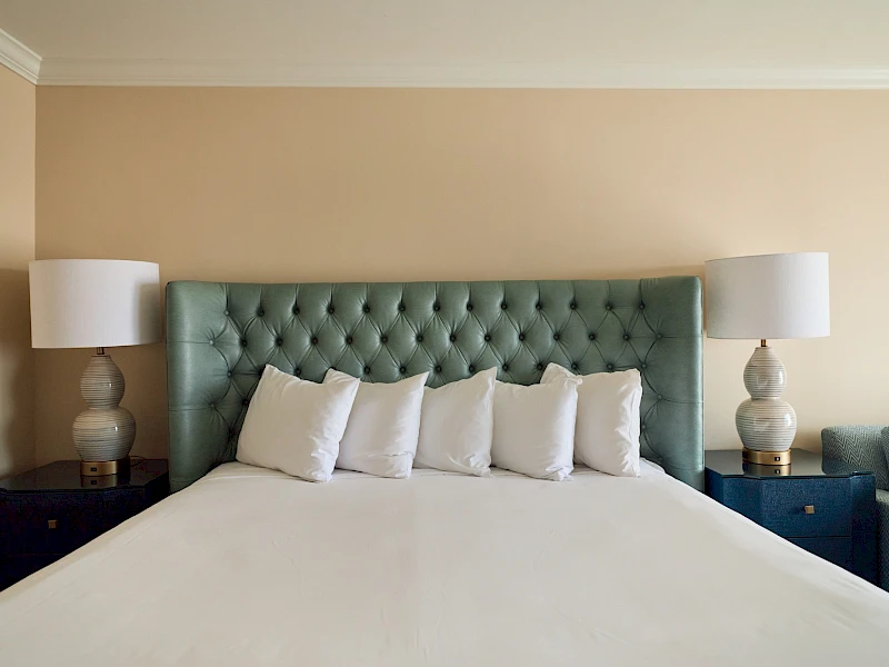 A neatly made bed with a tufted green headboard, six white pillows, cream walls, and matching nightstands with lamps on either side.