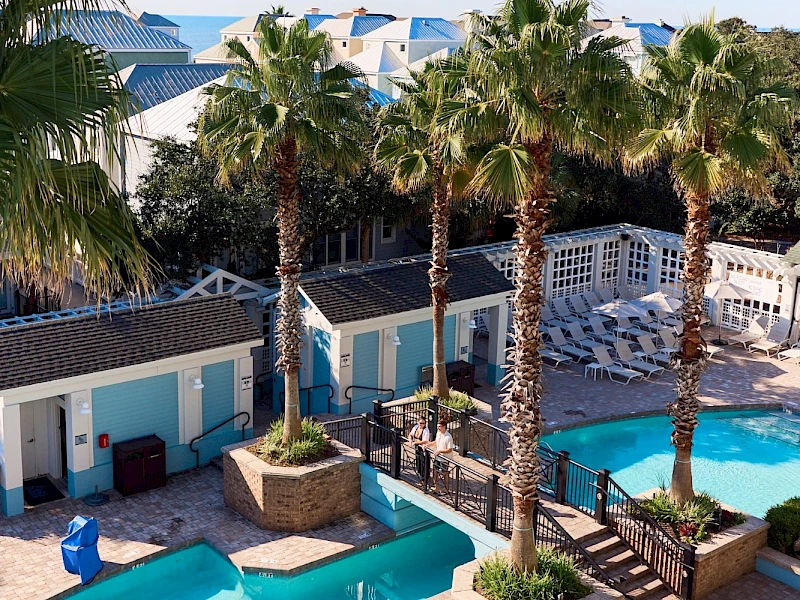 A sunny resort pool area with turquoise waters, palm trees, blue and white cabanas, and shaded lounge spots by the deck, perfect for relaxing.