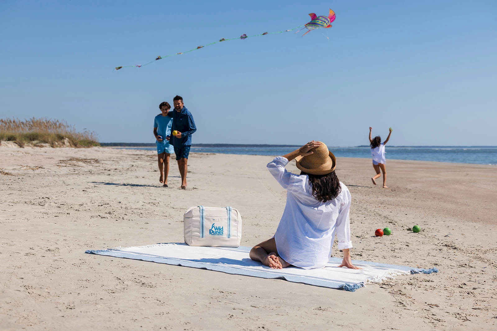 A woman sits on a blanket at the beach taking a photo of a kite in the sky, while two friends stroll nearby and a man flies a kite in the distance.